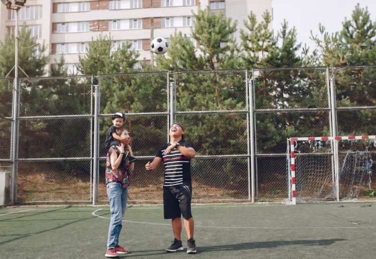 Full body cheerful father playing with football ball and mother giving piggyback ride to cute son while walking together on sports ground