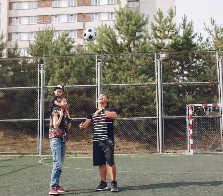 Full body cheerful father playing with football ball and mother giving piggyback ride to cute son while walking together on sports ground